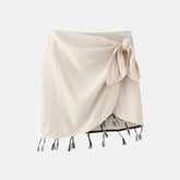 Saia curta bege com design envolvente, amarração lateral formando um laço e acabamento com detalhes de franjas pretas e brancas na barra, ideal para um visual boho ou casual.